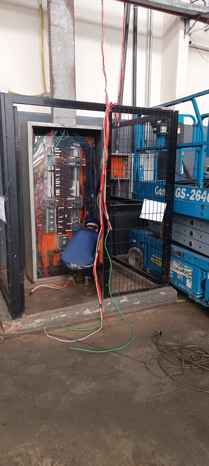 Painel elétrico aberto com circuitos e cabeamento em montagem, ao lado de plataforma elevatória.