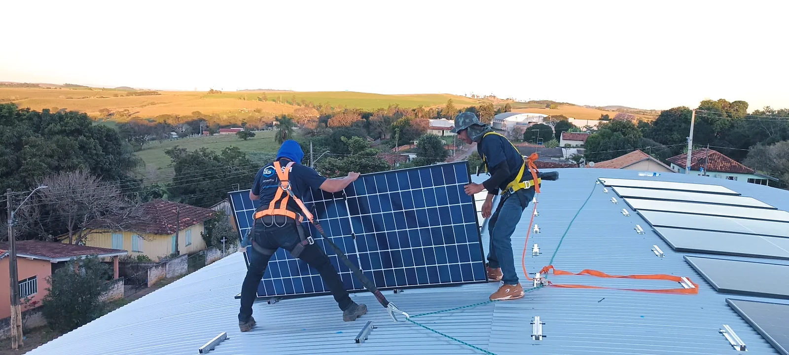 Técnicos com cintos e linha de vida instalando módulos fotovoltaicos em cobertura metálica.